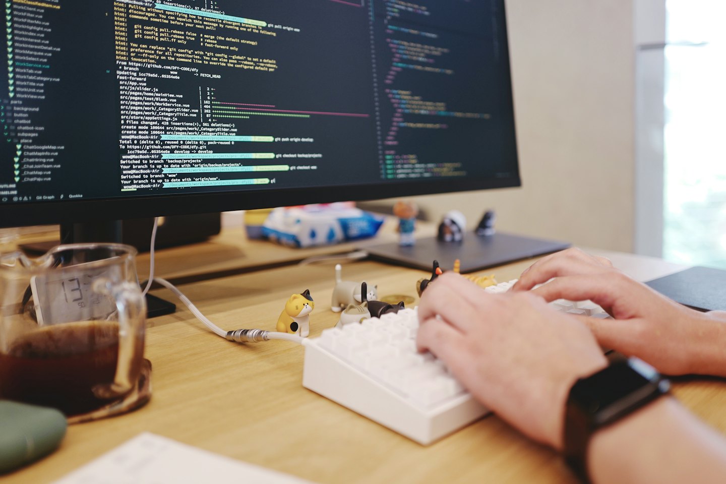 Hands type code on a keyboard near cat figurines and coffee.